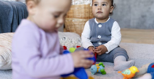 two toddlers sit on the floor separately holding toys. One is in focus in the background and the other in the foreground is out of focus.